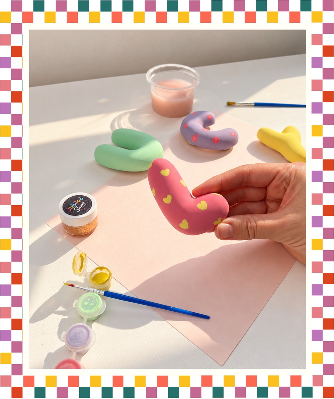 A hand holding a painted plaster bubble letter beside pastel letters, paint pots and brushes on a bright desk